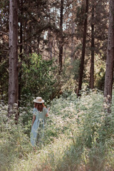 Portrait of a dreamy and relaxed happy red-haired girl in a linen rustic dress and a straw hat in a pine grove surrounded by white wildflowers. Walking in forest