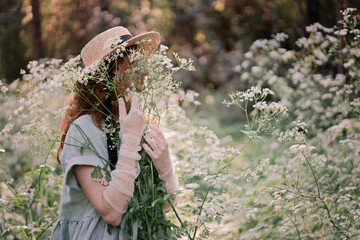 Portrait of a dreamy and relaxed happy red-haired girl in a linen rustic dress and a straw hat in a pine grove surrounded by white wildflowers. Walking in forest