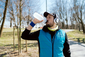 A man wearing wireless headphones drinks water from a reusable bottle. Athlete jogs in park in morning, jogging, taking care of health, figure, proper hydration of the body.