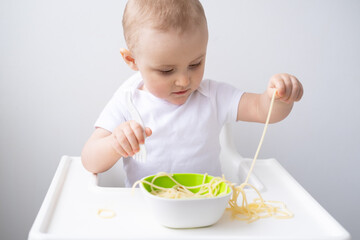 cute baby girl eating spaghetti pasta sitting in baby chair on white kitchen