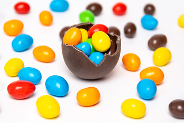 Half of a cracked chocolate egg filled with multi-colored candies on a white background with multi-colored candies, top view. Treats for children. Sweet dessert for kids. Sweet Easter tradition.