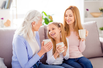 Photo of three cheerful idyllic people sitting couch hold drink mug communicate house indoors