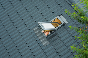 Little cheerful boy looks up into an open skylight window on roof of house covered with metallic...