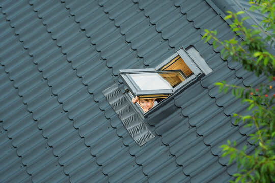 Little cheerful boy looks up into an open skylight window on roof of house covered with metallic gray tiles. Child breathes clean air. Summer morning. Top view.