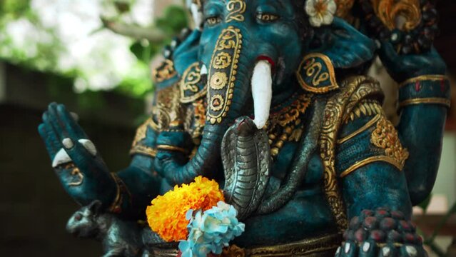 An old stone statue of Ganesha covered with moss with a wreath of yellow flowers on his neck standing on the street with green plants