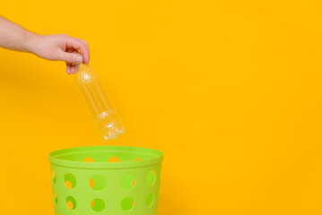 A man's hand throws a plastic bottle out of the water into a trash can for disposal and recycling. Environmental protection concept, yellow background, space for text.