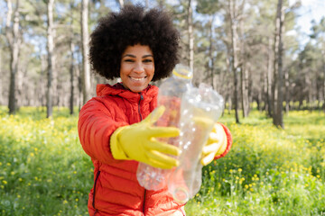 Smiling volunteer with protective glove holding a plastic bottle in the forest - African American woman caring for the environment.