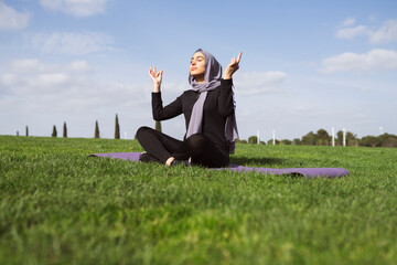 Happy young Muslim woman doing yoga exercises on green park background. Healthy lifestyle and good wellness concepts.