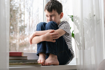 A sad boy sits on the windowsill hugging his knees. Bad mood, depression. The boy is sad alone at home