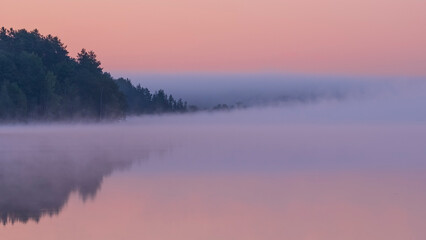 Naklejka na ściany i meble Dawn over the foggy lake. Beautiful dreamy view. Pink sky just before the sunrise and fog over water and trees with reflections on river bank.