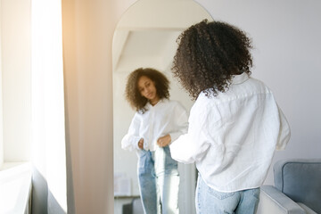 Cute african american girl choosing clothes to wear in front of mirror in i the living room....