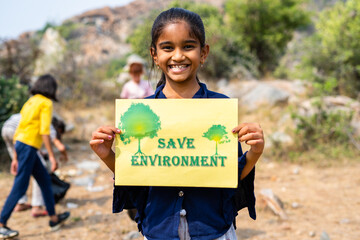 Happy smilig kid holding save environment sign board while other kids cleaning around at hill top - concept of support for climate change, sustainable lifestyle and volunteers