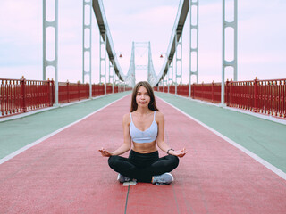 beautiful, sporty, fit, young caucasian woman in sportswear and sneakers sitting in lotus pose on the bridge. Sport, activity, yoga, urban lifestyle concept