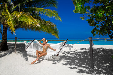 Beautiful sexy tanned woman in golden bikini on Maldives island. Young glamour girl is posing on hammock in paradise . Perfect body bikini model on Maldives beach. Bikini fashion. Luxury travel.