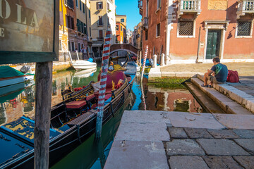 VENICE, ITALY - August 27, 2021: View of empty gondola on narrow canals, with a tourist waiting for the gondolier of Venice, Italy.