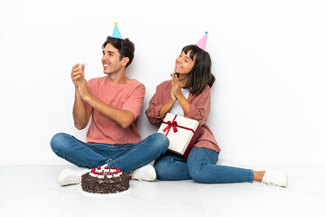 Young mixed race couple celebrating a birthday sitting on the floor isolated on white background applauding after presentation in a conference