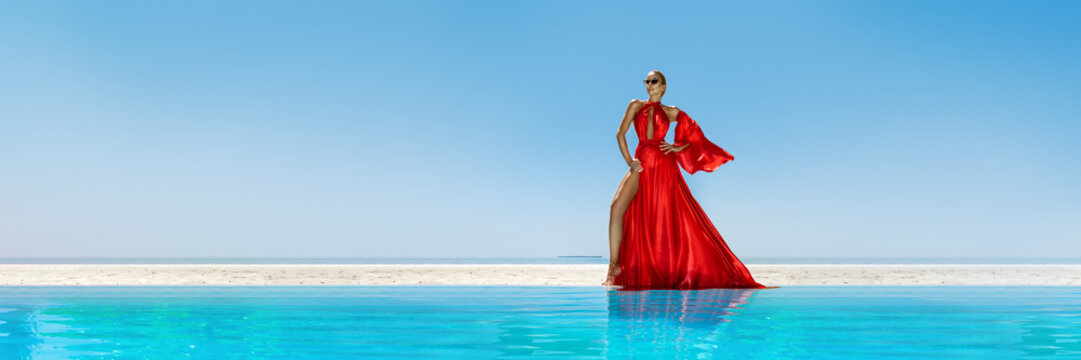 Luxury fashion. Elegant fashion model. Stylish female model in red long gown dress on the Maldives beach. Elegance. Classy woman in amazing red dress near the pool. Couture. Vogue. Panorama.