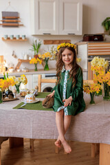 Happy Easter! cheerful beautiful girl in a green dress with a flower wreath plays with a rabbit at home in the kitchen