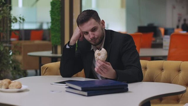 Young sad Caucasian businessman sitting at the table and eating donut. Portrait of stressed handsome man having break indoors. Lifestyle and business concept