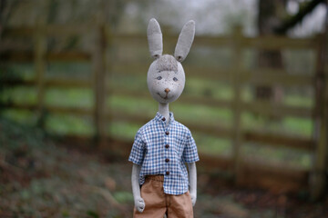 Сute doll rabbit in a  plaid shirt standing in front of the wooden fence.
