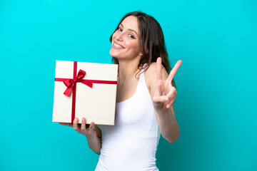 Young caucasian woman holding a gift isolated on blue background smiling and showing victory sign