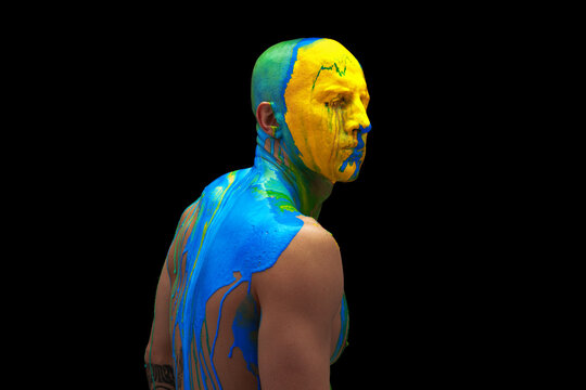 Portrait of young man with closed eyes being covered with thick multicolored paint on his head and shoulders isolated over black background