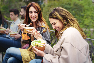 Gathering of young people eating salads in plastic-free bowls, talking, joking and having fun together sitting outdoors in the park - obrazy, fototapety, plakaty