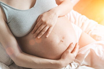 A pregnant woman strokes her belly with a baby before childbirth while relaxing in a sunny bright bedroom lying on a bed. Women's health, pregnancy, conception, childbirth concept.