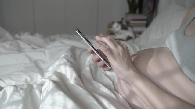 A pregnant woman uses the phone and strokes her belly while relaxing in a home bed. Using a smartphone before the birth of a child. Women's health, remote consultation with a doctor, pregnancy period