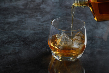 Pouring brandy alcohol drinks into the glass of brandy with ice cubes and reflective on black counter bar background.
