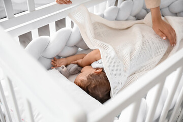 Young mother covering her sleeping baby with blanket in crib at home