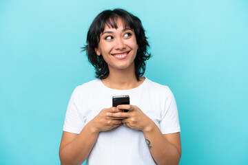 Fototapeta premium Young Argentinian woman isolated on blue background using mobile phone and looking up