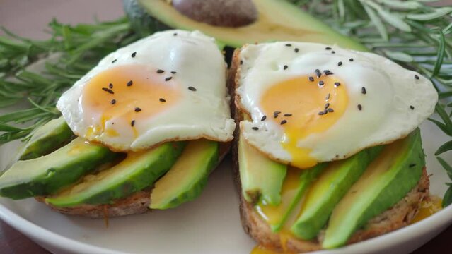 Close up Two Toasts with avocado and fried egg Spreading Yolk, rosemary 4K. Preparing healthy toast breakfast avocado slices rye wholegrain bread, sunny side up egg on top. breakfast for two, couple