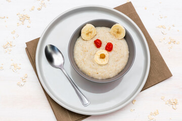 Funny cute kids childrens baby's healthy breakfast lunch oatmeal porridge in bowl look like bear face decorated with banana, dried berry fruits. dessert food art on white wooden table