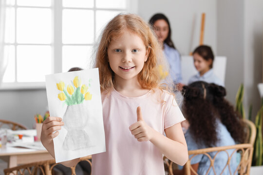 Cute redhead girl showing thumb-up and holding picture painted during master-class in art