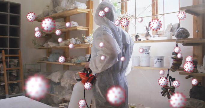 Image of covid 19 cells over person in ppe suit and face mask disinfecting pottery studio