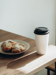 Cup of coffee to go in white paper cup and croissants in sunlight on wood table.