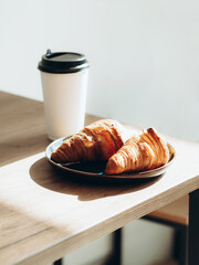 Cup of coffee to go in white paper cup and croissants in sunlight on wood table.