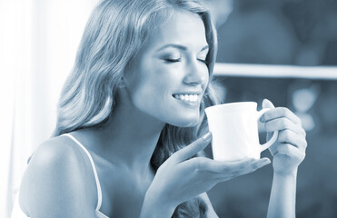 Blue tone monochrome image of young happy smiling woman with white cup near window, at home. Cheerful amazed blond girl with closed eyes drinking coffee indoors. Morning calm concept.
