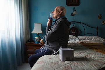 Lateral view angle portrait of an old man with asthma using a nebulizer in his room sitting on a bed - obrazy, fototapety, plakaty