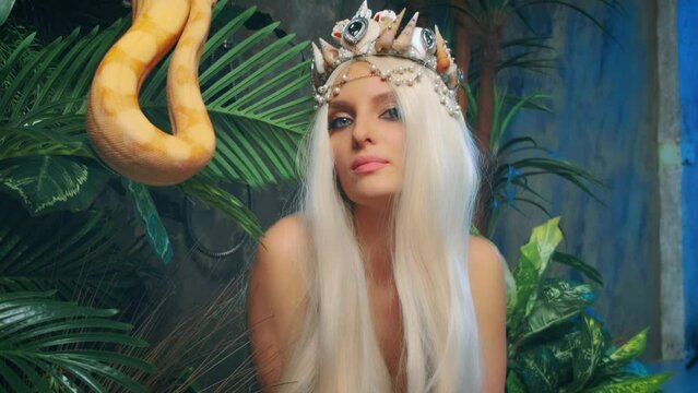 Close-up art portrait woman with snake. Nagga goddess touches white milk Boa constrictor. Fantasy asp girl blonde long hair, looks at serpent. Lady goth vampire queen viper. Backdrop green palants. 4k