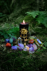 Black candle with sun amulet, mineral gemstones, flowers in forest, dark natural forest background....