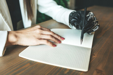 A close-up photo of a young disabled girl with a bionic prosthesis writes in a notebook, sitting at a table. Online work, distance learning. 