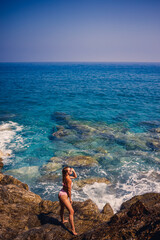 Young beautiful woman in a swimsuit stands on a rocky beach of the Mediterranean Sea. The concept of sea recreation. Selective focus