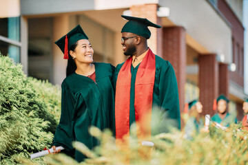 Happy Asian graduate and her black classmate talk on their graduation day.