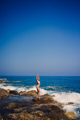Beautiful young woman in a swimsuit on a rocky beach on a sunny day against the backdrop of waves. Vacation in the summer season. Selective focus