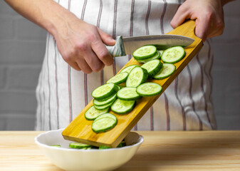 A young man puts a sliced cucumber in a plate for making a salad.