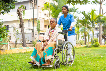 wide shot of Nurse taking senior woman on walk while on wheelchair at hospital garden - concept of caretaker, disability and healthcare