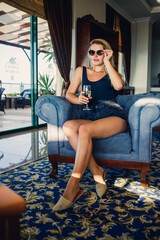 Obraz premium Beautiful young woman sits on a soft armchair in the hotel lobby with a glass of champagne on vacation. She is wearing sunglasses and a smile on her face. Vacation concept
