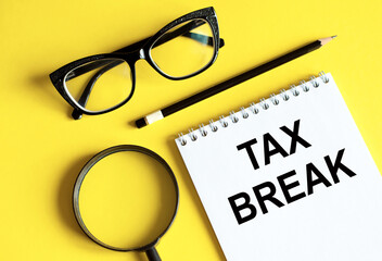 The phrase Tax Break in notebook with pencil, magnifier, and glasses on a yellow background